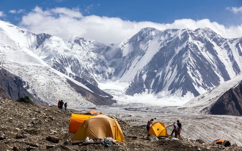 Vu de loin, deux tentes oranges installées et un duo de personne installe une autre tente, aux pieds des montagnes enneigées.