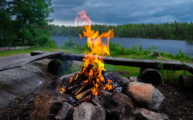 Feu de camp entre de grosses pierres et des rondins et planches forment des bancs autour du feu. On voit en arrière-plan un lac et une foret.