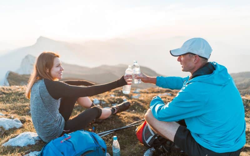 Une femme et un homme assis dans la nature face à des montagnes au loin, trinquant avec deux bouteilles d'eau, un sac de randonnée est posé à côté d'eux.