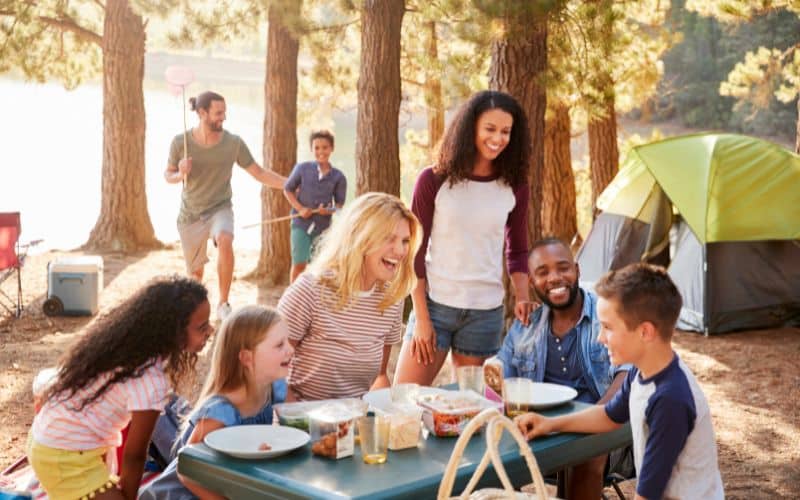 Famille assise à une table de camping avec adultes et enfants partageant un bon moment, tous ris et ont de quoi manger devant eux. On voit une tente en arrière-plan.